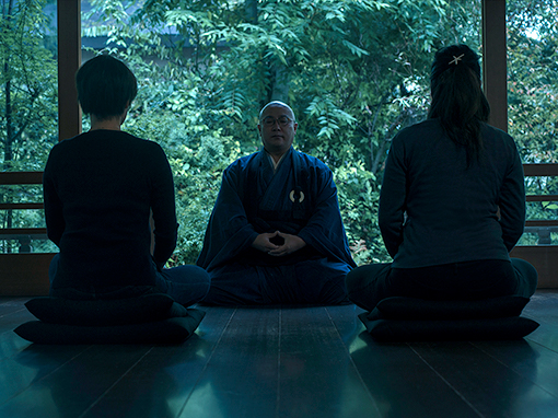 Two guests practising zazen with an instructor in a room overlooking greenery